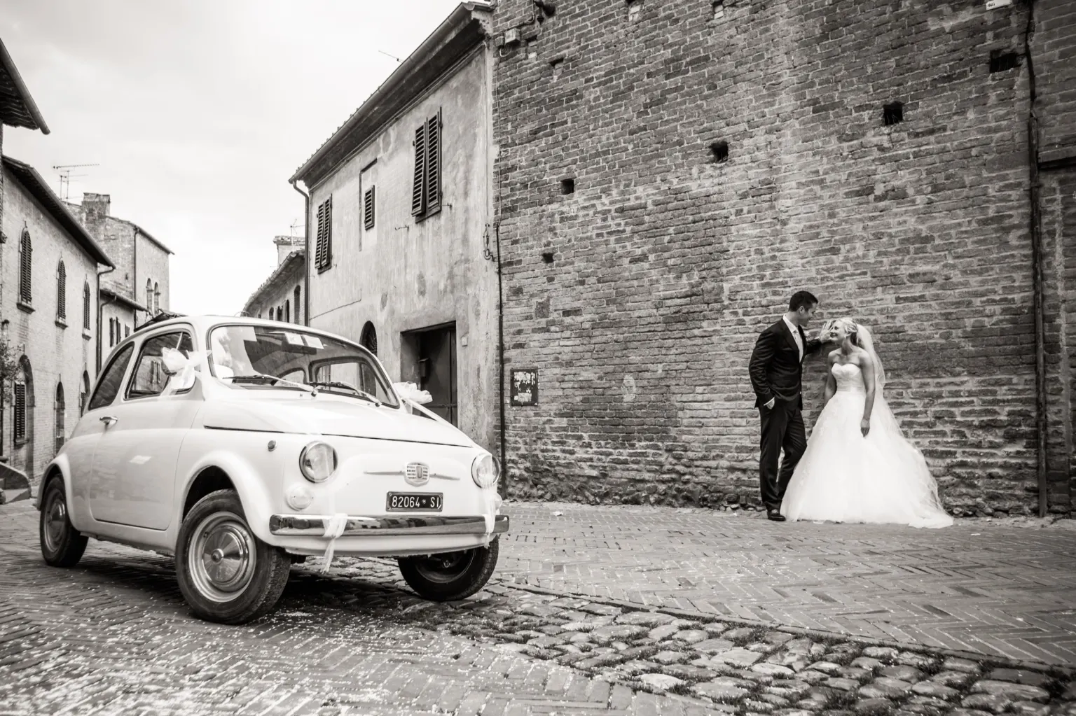 Bride and groom kissing next to a vintage Fiat 500 in a Tuscan village street