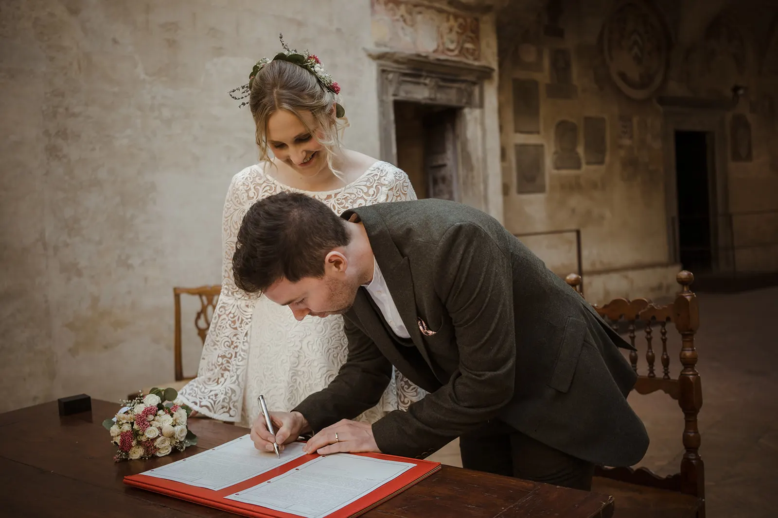 Efffetti® Wedding Planners In Tuscany - Italy Groom signing the marriage register at a civil wedding in Certaldo, the bride smiling behind him