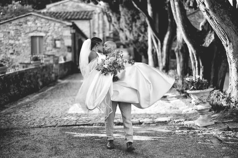 Black and white photo of the groom carrying the bride and kissing her on a cypress-lined path in Tuscany, photo Daniele Vertelli