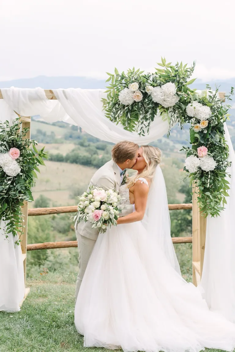 Bride and groom kissing under a flower arch overlooking the Tuscan hills