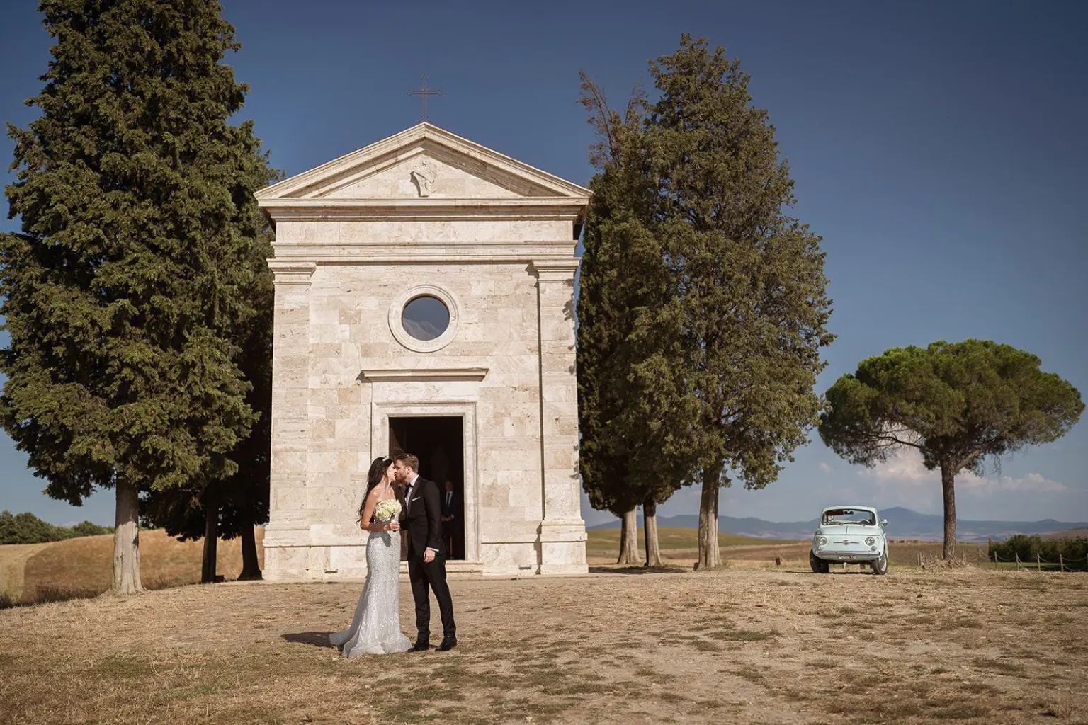 The chapel of the Bride and groom kissing in front of the chapel of the Madonna di Vitaleta, with a vintage Fiat 500 among the cypresses